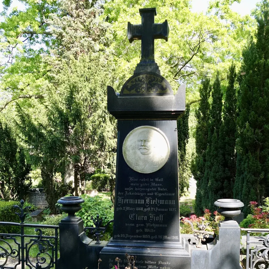 Schwarzes Grabmal mit Relief des Bestatteten und altdeutscher Schrift. Auf dem Grabstein ein großes Kreuz. Dahinter: Beete mit bunten Blumen und grüne Bäume.