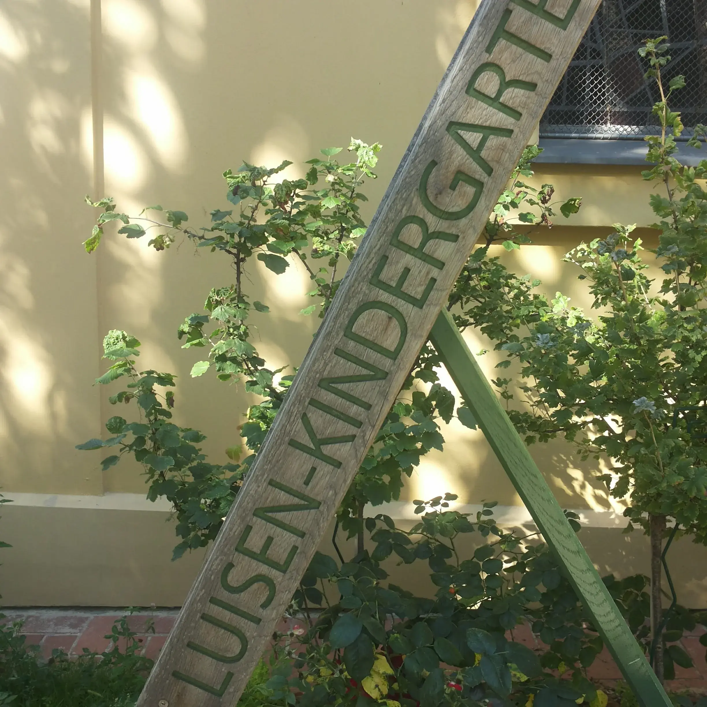 Hölzernes Schild mit der Aufschrift "Luisen-Kirchengarten" vor der gelbem Kirchenfassade und Rosenbüschen.