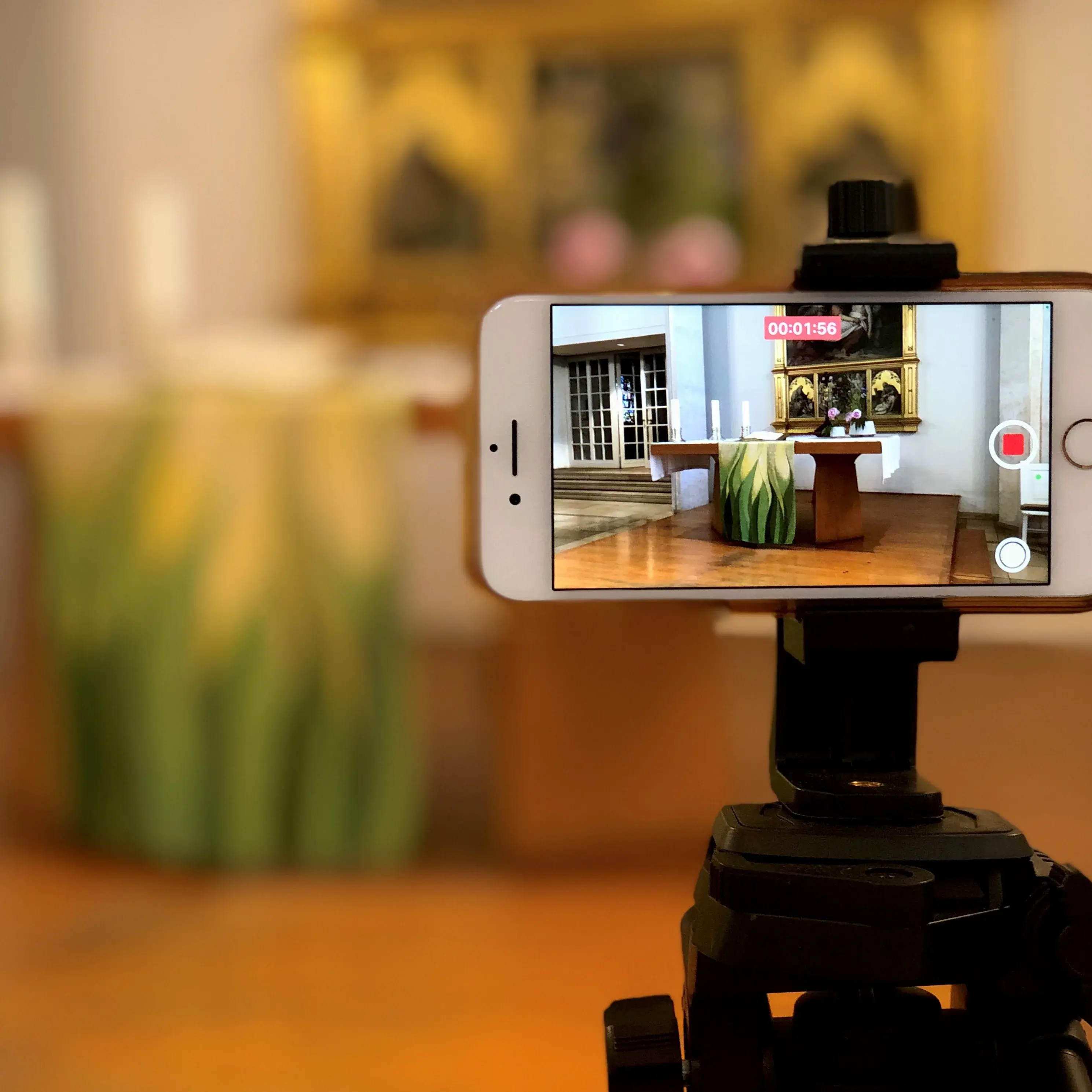 Stativ mit Smartphone, das den Altar filmt. Der Altarraum ist im Hintergrund unscharf zu erkennen. Auf dem Handybildschirm ist er scharf zu erkennen: Holzboden, Holz-Altar mit Kerzen, Blumen und einem grün-gelbem Altartuch. Dahinter Altarbild im goldenen Rahmen mit Kreuzigungsszenen.