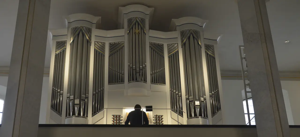 Orgelempore der Luisenkirche mit weißer Orgel und silber-glänzenden Orgelpfeifen. Kantor Jack Day ist von hinten zu sehen, wie er an der Orgel spielt.
