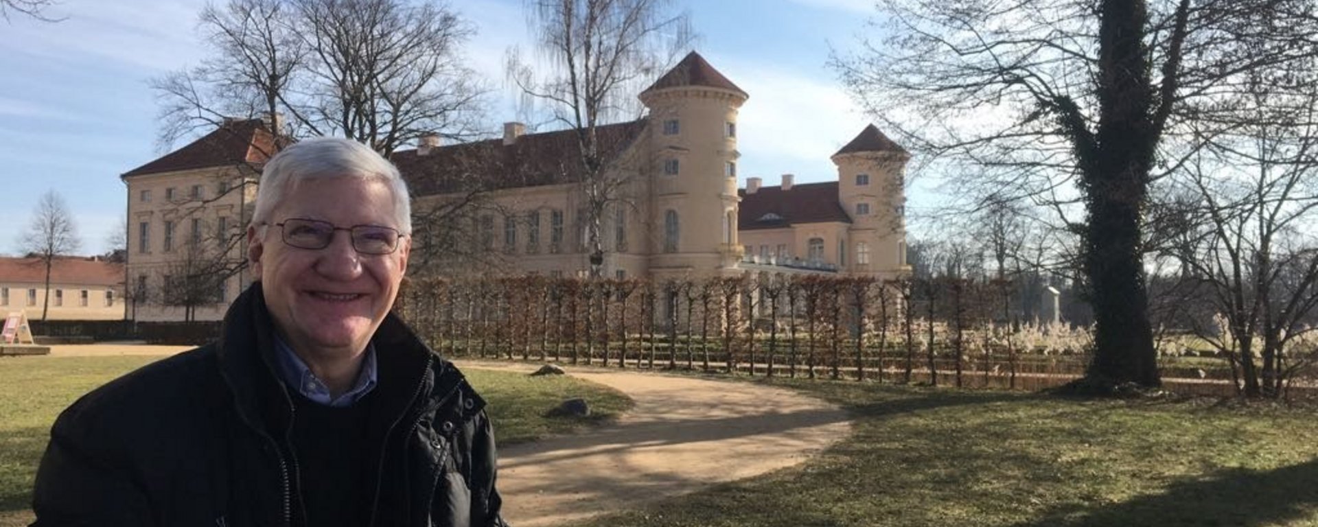 Raymond Hughes (schwarze Jacke, schwarzer Pullover, blauer Hemdkragen, graue kurze Haare, Brille, freundliches Lächeln) in einem Park mit Schloss im Hintergrund