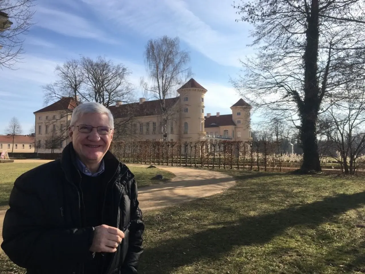 Raymond Hughes (schwarze Jacke, schwarzer Pullover, blauer Hemdkragen, graue kurze Haare, Brille, freundliches Lächeln) in einem Park mit Schloss im Hintergrund