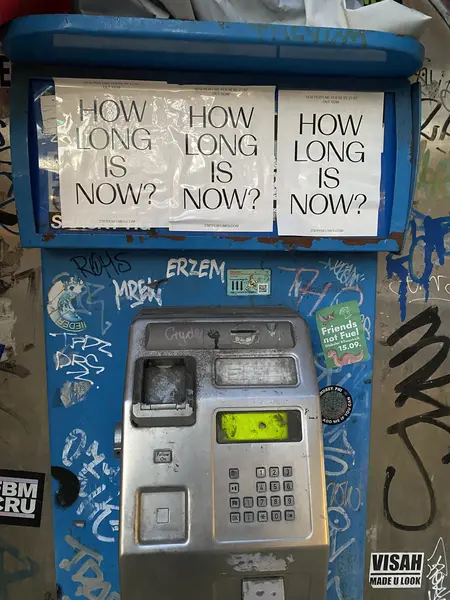 Foto einer bemalten blauen Telefonzelle mit demoliertem Telefon. Über dem Telefon drei Aufkleber mit der Aufschrift &quot;HOW LONG IS NOW?&quot;