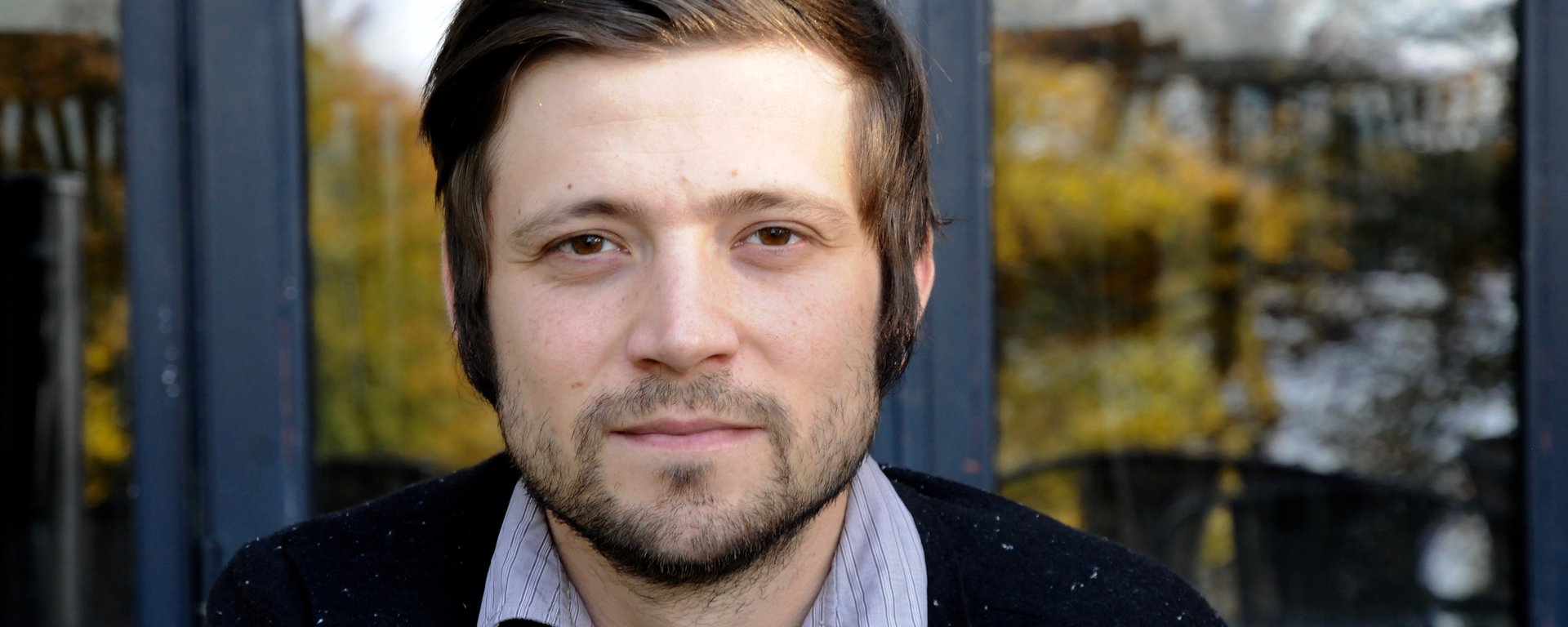 Portraitfoto von Alexandru Bulucz vor einem Fensterrahmen, in dem sich grüne Bäume spiegeln. Bulucz trägt ein schwarzes Jacket und ein blau gestreiftes Hemd (man sieht nur den Kragen), braune Haare, nach rechts gekämmt und Koteletten, Drei-Tage-Bart, braune Augen.