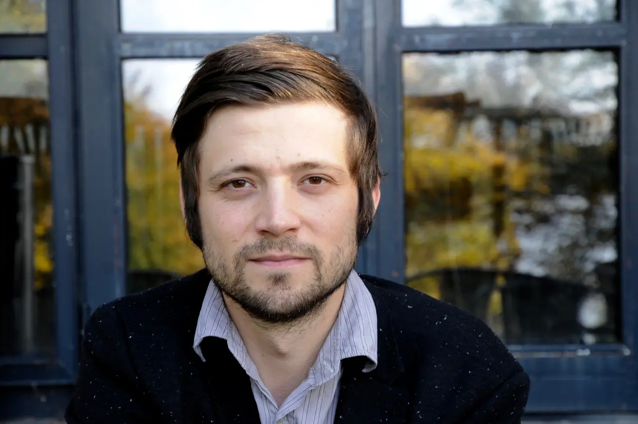 Portraitfoto von Alexandru Bulucz vor einem Fensterrahmen, in dem sich grüne Bäume spiegeln. Bulucz trägt ein schwarzes Jacket und ein blau gestreiftes Hemd (man sieht nur den Kragen), braune Haare, nach rechts gekämmt und Koteletten, Drei-Tage-Bart, braune Augen.
