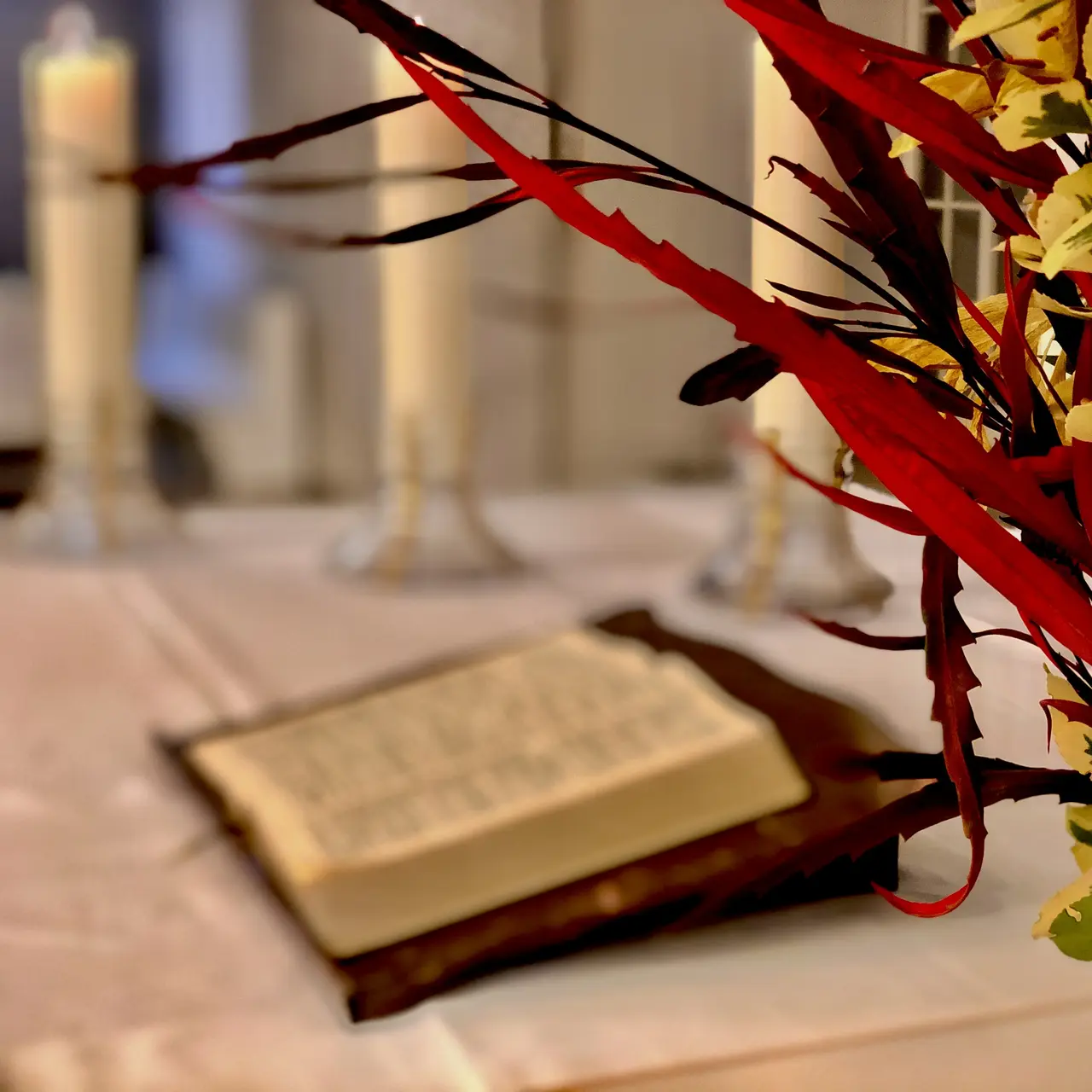 aufgeschlagene Bibel auf dem Altar, der mit einem weißen Tuch bedeckt ist. Ebenfalls auf dem Altar sind - umscharf - im Vordergrund ein Strauß bunter Blumen und im Hintergrund drei Kerzen gezeigt.