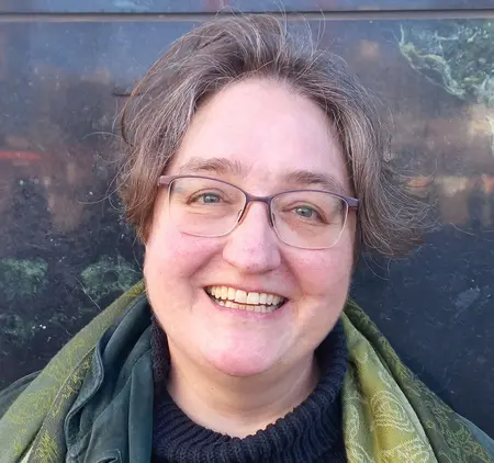 Porträt von Pfarrerin Anne Hensel. Dunkler Pulli mit grünem Tuch. Rundes Gesicht, freundliches Lachen, Brille, kurze dunkle Haare.